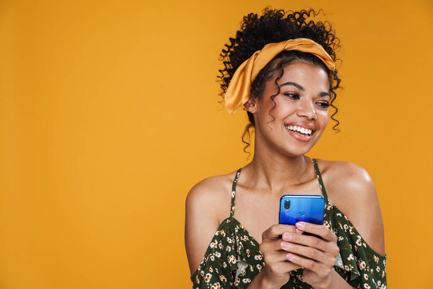 Image of happy african american woman smiling and using cellphone isolated over yellow wall - Photo, Image