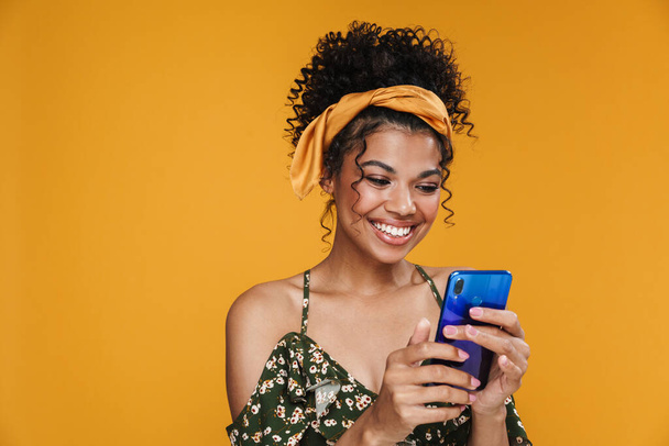 Image of happy african american woman smiling and using cellphone isolated over yellow wall - Foto, immagini