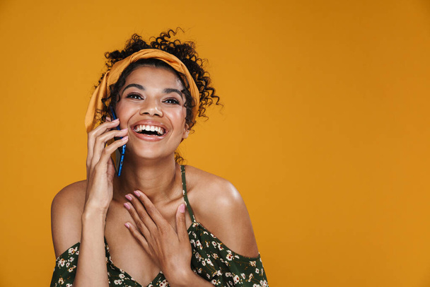 Beeld van gelukkige Afrikaanse Amerikaanse vrouw glimlachend en pratend op mobiele telefoon geïsoleerd over gele muur - Foto, afbeelding
