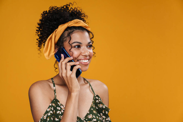 A boldog afro-amerikai nő képe, amint mosolyog és beszél a sárga falon izolált mobiltelefonján. - Fotó, kép