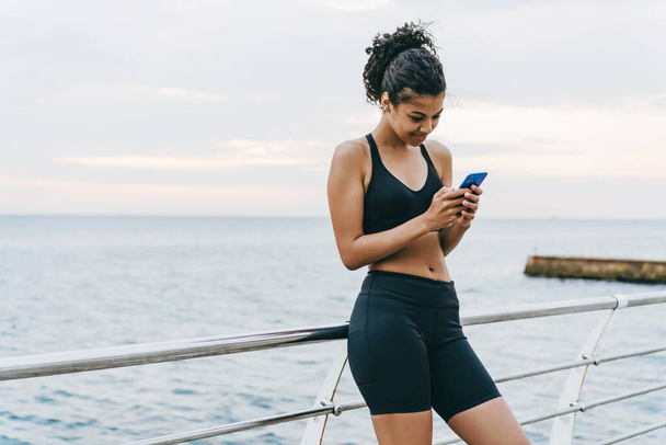 Imagen de la deportista afroamericana complacida usando teléfono celular en el paseo marítimo - Foto, Imagen
