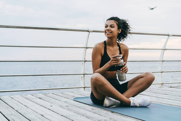 Image of smiling african american sportswoman drinking water while sitting on mat at promenade - Foto, immagini