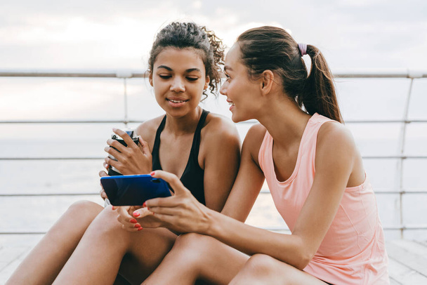 Imagen de deportistas multinacionales sonrientes usando teléfono celular mientras están sentadas en el paseo marítimo - Foto, Imagen