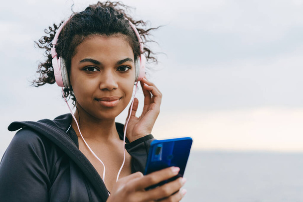 Image of pleased african american woman using cellphone and headphones while walking on promenade - Φωτογραφία, εικόνα