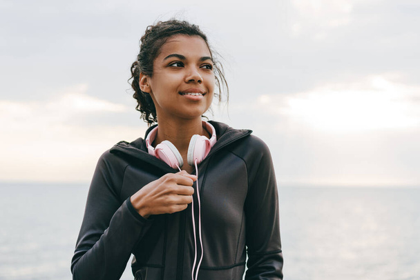 Image of young african american woman with headphones smiling while walking on promenade - Photo, image