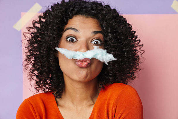 Image of amusing african american woman making fun cotton with candy isolated over multicolored background - Photo, image