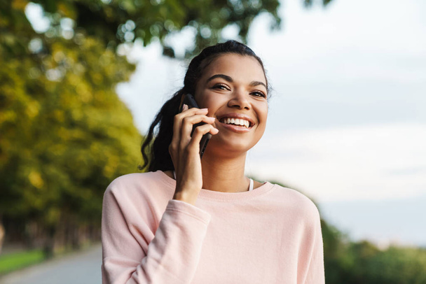 Image of happy african american girl talking on mobile phone while walking in summer park - Fotó, kép