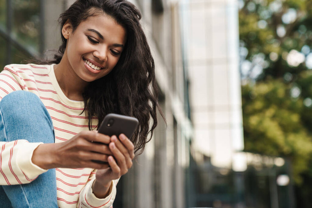 Image of smiling african american girl using mobile phone while sitting at city street - Foto, Bild