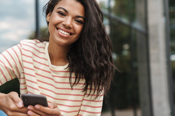 Image of smiling african american girl using mobile phone while sitting at city street - Photo, Image