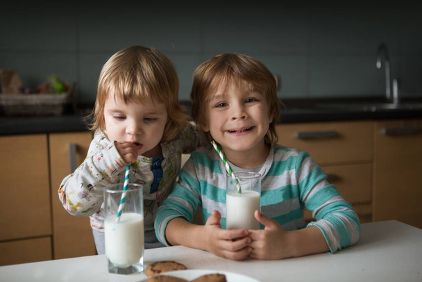 A little cute boy and his little sister drink milk and eating cookies in kitchen. - Fotó, kép