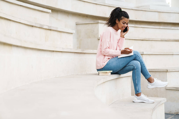 Image of smiling african american student girl talking on cellphone while sitting on stairs outdoors - Фото, изображение