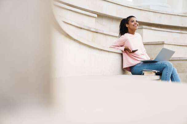 Image of joyful african american student girl using laptop and cellphone while sitting on stairs outdoors - Valokuva, kuva