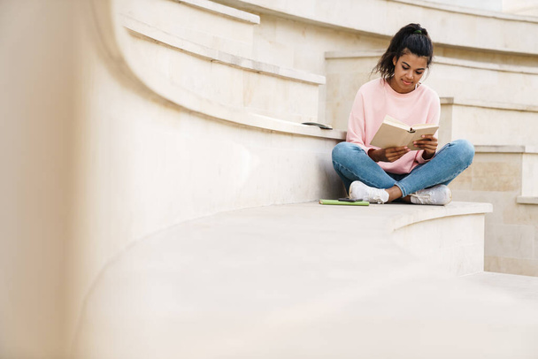 Image of focused african american student girl reading book while sitting on stairs outdoors - Fotografie, Obrázek