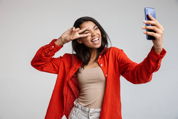 Young funny african casual lady grimacing and making selfie on smartphone isolated over gray, showing peace gesture - Foto, Bild