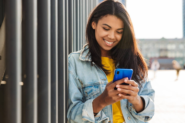 Image of joyful african american woman smiling and using smartphone while standing at city street - Photo, image