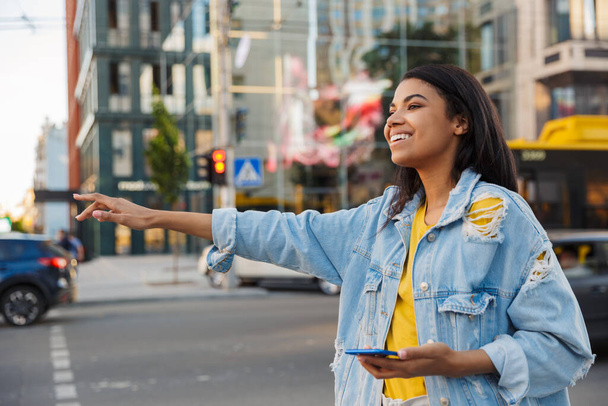 Attractive young african woman in a city street waving for taxi, holding mobile phone - Foto, immagini
