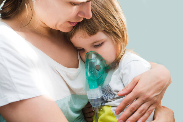 Little child girl inhaling from the inhaler with nebulizer at home. A child  with respiratory disease  in a mask for inhalations sits on hands at the mother. Asthma patien - Foto, Bild
