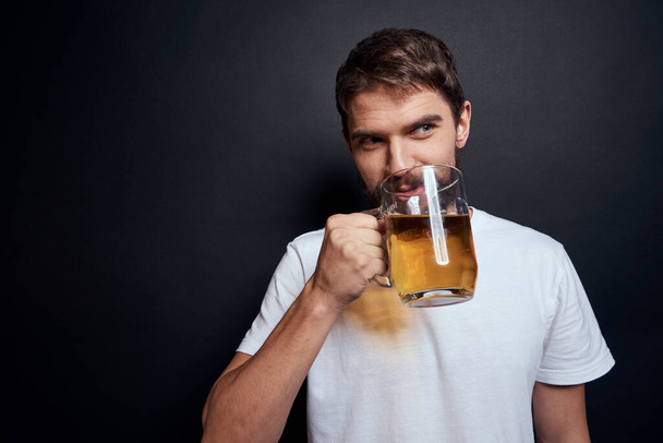 白いTシャツを着たビールのマグカップを手にした男は暗い隔離された背景で酒を飲み - 写真・画像