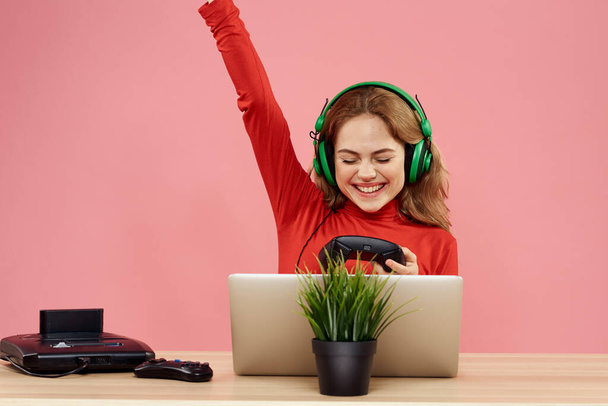 woman in headphones with gamepad in front of laptop sit at table entertainment lifestyle pink background - Foto, imagen