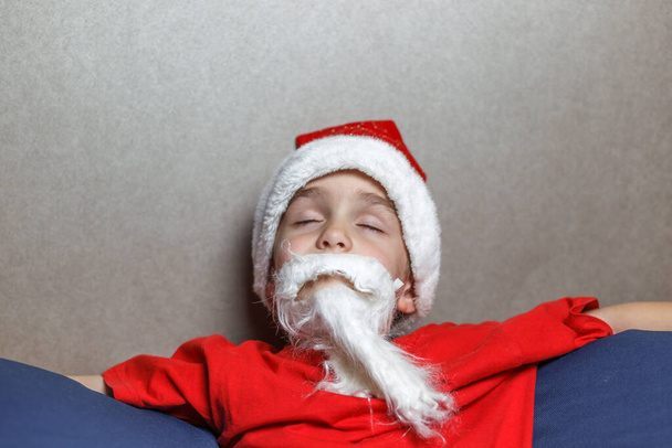 Ein Junge im Weihnachtsmannanzug und künstlichem Bart sitzt mit geschlossenen Augen auf einem Sackstuhl. - Foto, Bild