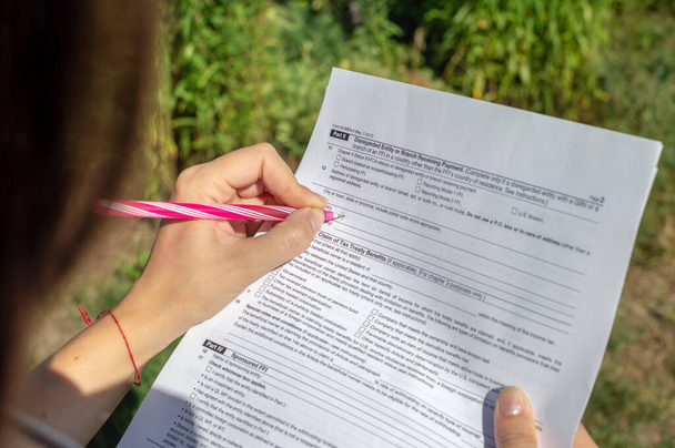 若い女性が納税申告書を手に取り、データを書き込む。芝生を背景にした野外活動 - 写真・画像