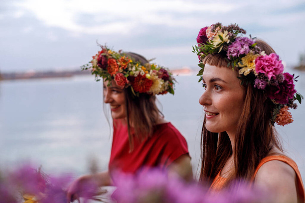 Δύο νεαρά και όμορφα κορίτσια με στεφάνια λουλουδιών στη φύση. Αρχαία ειδωλολατρική αντίληψη εορτασμού προέλευσης. Μέρα θερινού ηλιοστασίου. Στα μέσα του καλοκαιριού. - Φωτογραφία, εικόνα