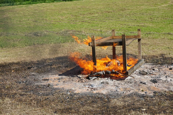 An old wooden table burns by the fire in the meadow during the summer.burning furniture - Photo, Image