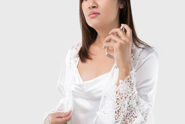 Uma mulher de camisola de cetim branco polvilhe perfume em si mesma e na garganta, em um fundo cinza claro - Foto, Imagem