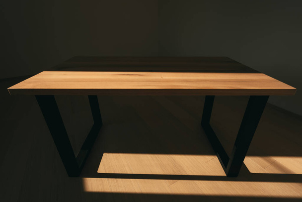 Mesa de madera moderna hecha de madera maciza y estructuras angulares de hierro en forma de patas en los lados.Mesa está en habitación vacía con pared blanca y suelo de madera. La luz del sol cae sobre la mesa y crea una escena de contraste. - Foto, imagen