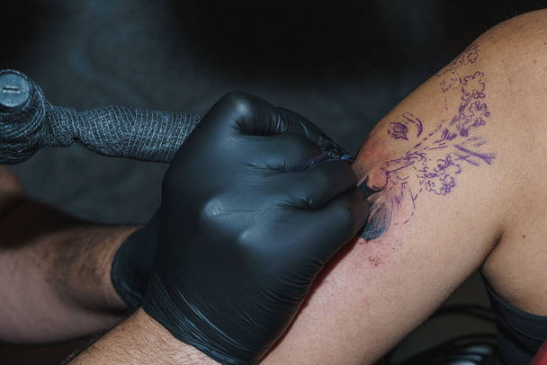 A nő karját egy COVID elleni védelemmel ellátott géppel tetoválják. Életstílus. Egy tetováló fekete latex kesztyűt visel. A tetoválás egy női arc.. - Fotó, kép