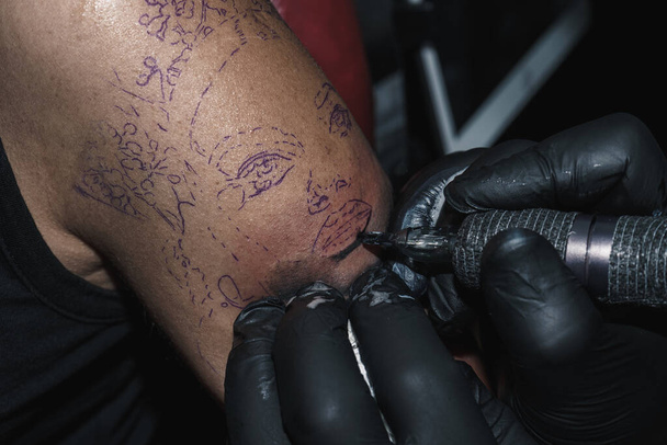 Woman's arm being tattooed with a machine protected against COVID. Lifestyle. Tattooist wearing black latex gloves. The tattoo is a woman face. - Photo, Image