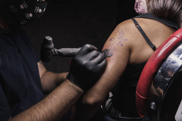 Naisen käsi on tatuoitu koneella, joka on suojattu COVID:ltä. Elämäntapa. Tatuoija mustissa lateksikäsineissä. Tatuointi on naisen kasvot. - Valokuva, kuva