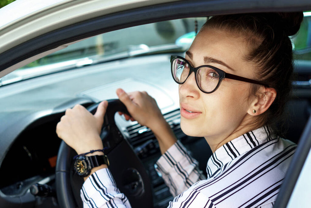 Geschäftsfrau mit schwarzer Brille und Hemd sitzt im Auto mit Lenkrad. Junges attraktives Chefmädchen im Business-Stil mit einer Armbanduhr eilt zur Arbeit. Beratender Makler im Salon - Foto, Bild