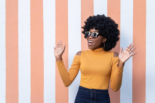Portrait of fashionable African happy woman with curly hairstyle, wearing sunglasses, raises palms, has glamour look, posing on striped background outdoors.  People and positive emotions concept. - Foto, Imagem
