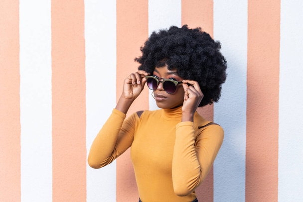Close-up portret van zelfverzekerde Afro-Amerikaanse elegante vrouw met krullend kapsel, het dragen van stijlvolle zonnebril, heeft fashion look, poseren op gestreepte achtergrond buiten. Lege kopieerruimte. - Foto, afbeelding