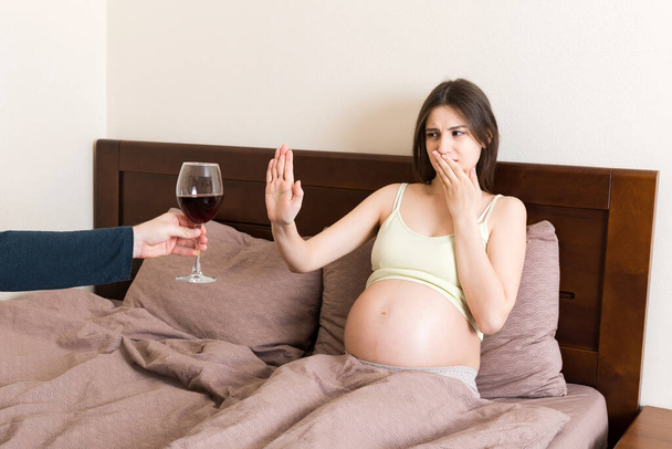 Pregnant woman staying in bed refuses to drink wine and makes stop gesture to the glass. No alcohol during pregnancy concept. - Photo, Image