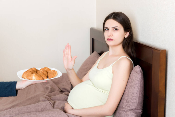 Pregnant woman relaxing in bed makes stop gesture to croissants. Expecting mother refuses to eat pastry. Diet during pregnancy concept. - Photo, Image