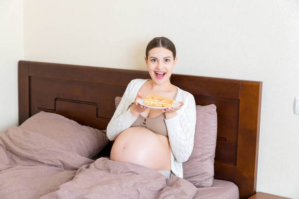 Голодна вагітна жінка їсть шматочок смачного торта, що розслабляється в ліжку вдома. Солодкий голод під час вагітності
. - Фото, зображення