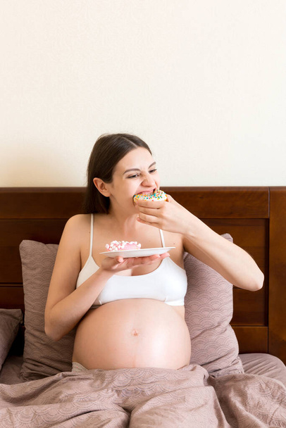 Pregnant woman is eating many donuts relaxing in bed. Unhealthy dieting during pregnancy concept. - Photo, image