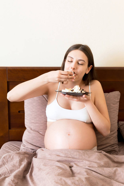 Hungry pregnant woman relaxing in bed is eating greedily a piece of cake and has a dirty mouth. Expecting mother can't stop eating. Strong appetite during pregnancy concept. - Photo, image