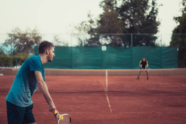 Πίσω όψη ενός άνδρα που κρατά μια ρακέτα και μια μπάλα, ετοιμάζεται να εξυπηρετήσει ενώ παίζει έναν αγώνα τένις έξω σε ένα χωράφι από πορτοκαλί πηλό - Φωτογραφία, εικόνα