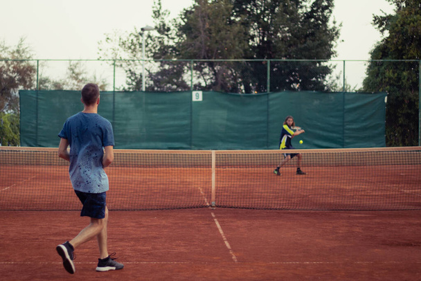 Hombre con camisa azul y pantalones azules visto desde atrás jugando al tenis, campo de juego naranja arcilla abierta. Anticipando el saque desde el otro lado, contorno del oponente golpeando una pelota - Foto, Imagen