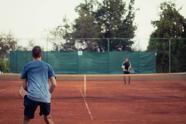 Homme en chemise bleue et pantalon bleu vu de derrière jouant au tennis, terrain de jeu orange argile grand ouvert. Anticipant le service de l'autre côté, contour de l'adversaire frappant une balle - Photo, image