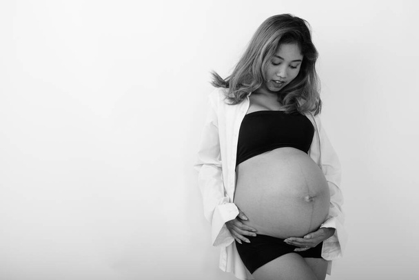 Studio shot της νεαρής εγκύου όμορφη Ασιάτισσα γυναίκα σε λευκό φόντο σε μαύρο και άσπρο - Φωτογραφία, εικόνα