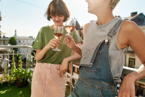 Cropped shot of two cheerful women holding glass of wine while having party, celebrating anniversary on the balcony. Young lesbian couple spending their day together - Photo, Image