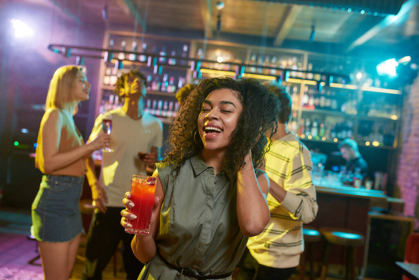 Attractive mixed race young woman smiling at camera while posing with a cocktail in her hand and friends chatting, having drinks at the bar counter in the background - Zdjęcie, obraz