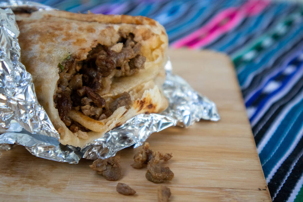 Carne Asada Burrito wrapped in foil, open with a bit taken out of it laying on a wooden board - Photo, Image