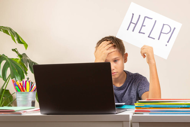 Trudności w nauce, szkoła, edukacja, koncepcja zdalnego uczenia się online. Smutny dzieciak siedzi przy stole z laptopem i trzyma kartę z Pomoc słowo - Zdjęcie, obraz