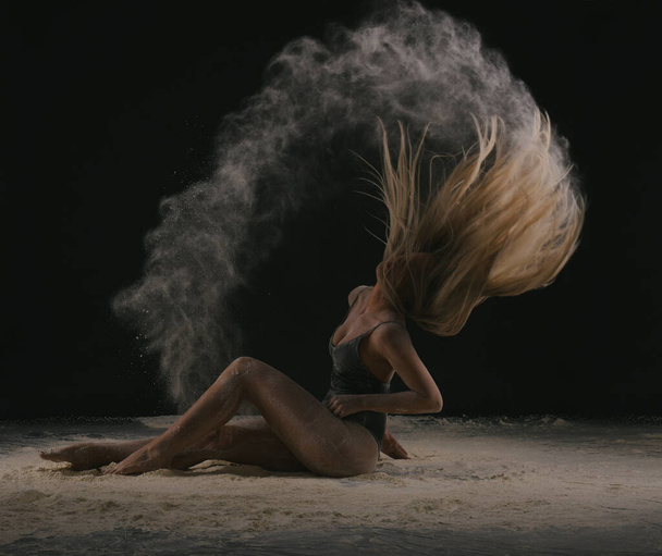Piękne zmysłowe dopasowanie szczupła tancerka w czarnym ciele z długimi blond włosami rzucanie pyłu, mąka, proszek na czarnym tle. - Zdjęcie, obraz