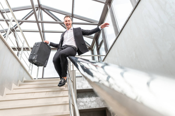 Empresário feliz vestindo terno formal deslizando em escadas Railing no edifício de negócios moderno. Conceito de pressa e liberdade - Foto, Imagem